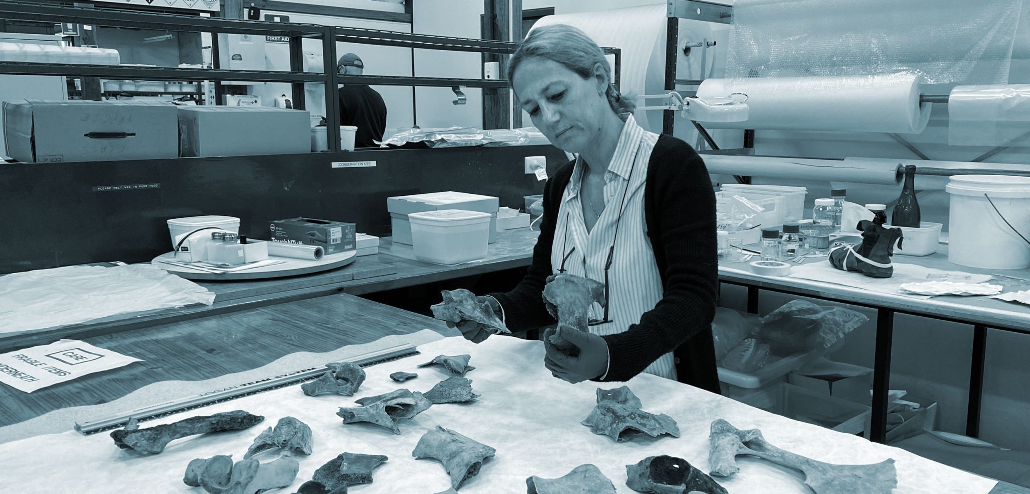 An black and white image of trowelblazer Edda Guareschi, a white woman with light colored hair wearing a striped button up shirt and a black cardigan with goggles hanging around her neck. She is wearing gloves and examining bone fragments in a lab space.