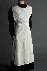 A smock-like white apron over a long-sleeved black dress on a mannequin.