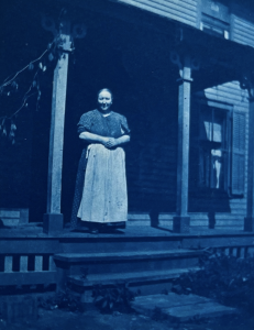 A cyanotype of a woman wearing a long white apron over a dark dress with sleeves rolled up. She is standing on the porch of a house.