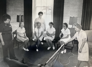 Black-and-white photo of seven women (six dressed in mid-length white dresses, one in matching dark-colored shirt and pants). Six women watch on while one vacuums.
