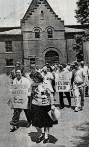 Black-and-white photo on newsprint of a group of people outside of College Hall carrying signs with slogans such as "show your support pro union pro smith" and "4.6% it's only fair"