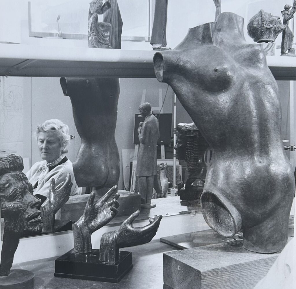 Black and white photo of Frances standing in her studio surrounded by many of her sculpture pieces