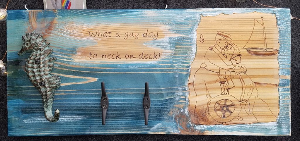Coat rack with wood burned line drawing of an early 1900s queer couple and wood burned phrase, "What a gay day to neck on deck"