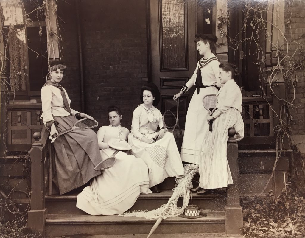 Photograph, black and white. Five women in long white dresses hold tennis equipment and sit on steps of a house.