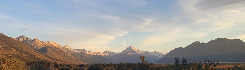 Landscape photo of Aoraki mountain located on the South Island on Aotearoa New Zealand. The morning sun shines golden on the rugged, snow capped peaks. The morning sky has a thin cloud layer with striking blue points. Short dark green shrubbery is in the foreground and moves outwards towards the mountains.