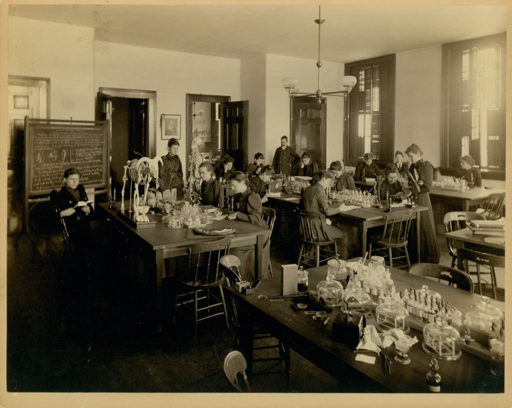 A wide view of a science lab. Women sitting at tables are working with books, animal skeletons, and beakers. A human skeleton in the background. Professor Wilder stands in the background.