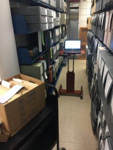 image of computer on a rolling cart in the middle of an aisle of archival boxes