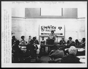 image of conference with women standing at podium in front of a banner that reads the elimination of racism