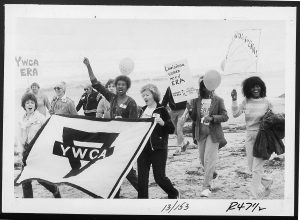 image of women marching on beach carrying banner for the YWCA and signs supporting the ERA
