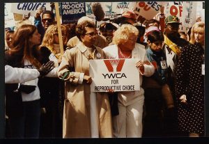 image of group of women marching for women's rights. Two women in the foreground hold a sign that reads "YWCA for reproductive rights"