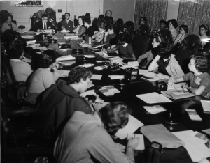 black and white photograph of women and girls sitting at a long table listening and taking notes 
