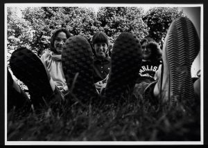 black and white photograph of three young women sitting in grass with leg in front of them. Feet are in the foreground.