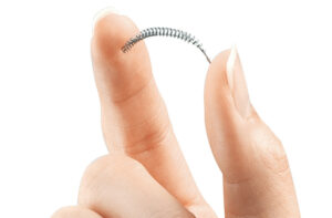 An Essure coil held between a white person's thumb and forefinger against a white background. The coil is a small, slightly curved silver coil