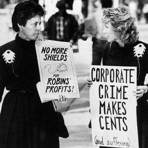 Black and white photo of women protesting against the Dalkon Shield, wearing large models on the shield on their shoulders. From left to right, the signs read "no more shields for Robins' profits!" and "corporate crimes makes cents (and suffering)"