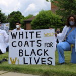 A white and black doctor kneeling with a poster that reads "white coats for black lives"