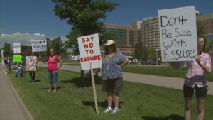 Photograph of protesters holding homemade signs outside of University of Colorado Hospital. From right to left, the first two signs read "don't be sure with Essure" and "say no to Essure"
