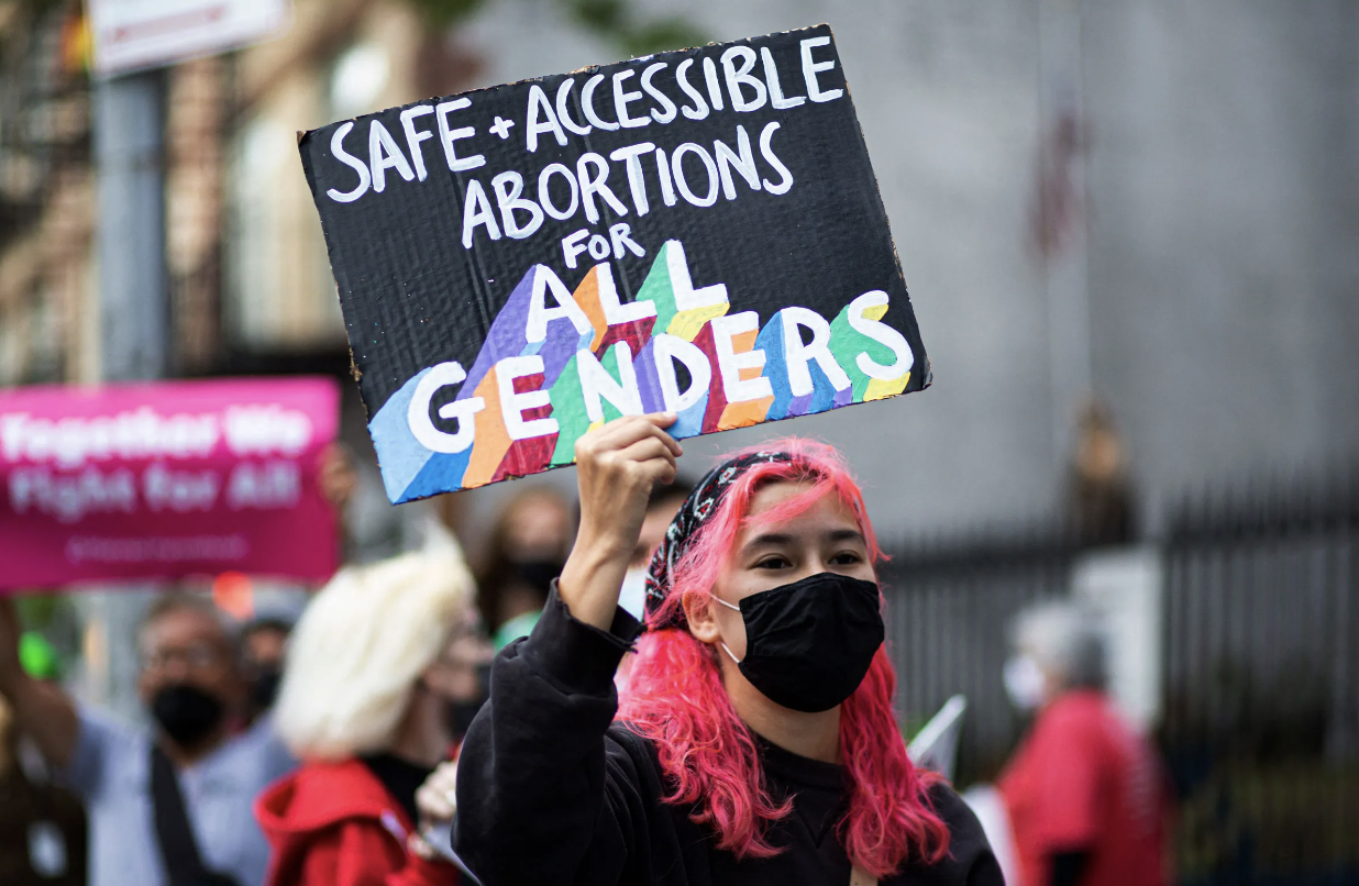 A masked protestor with pink hair holding up a sign that reads "Safe + Accessible Abortions for All Genders."