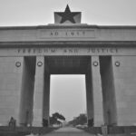 A black-and-white photo of an independence monument in Accra, Ghana. The monument is an arch with a black star on the top, above the date "AD 1957," the date Ghana gained independence, and then the words "Freedom and Justice."