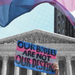 Photograph of a hand holding up a sign that reads "Our Bodies Are Not Our Destiny" with a transgender pride flag in the background.