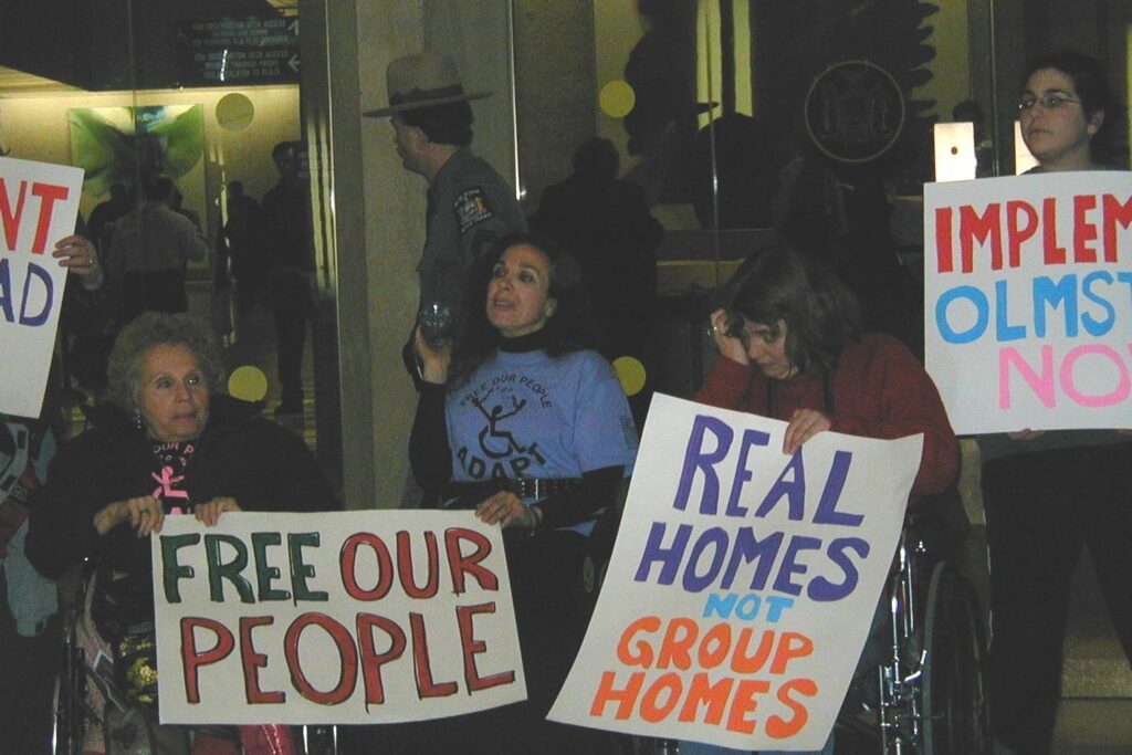 A group of people- all in various states of distress- are in wheelchairs and holding various signs that are protesting the Olmstead Law