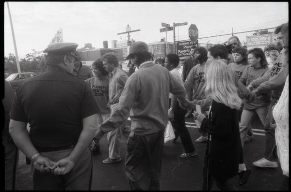 Photograph of escorts ushering a woman past police and pro-life protesters into the Providence Planned Parenthood clinic, 1989.