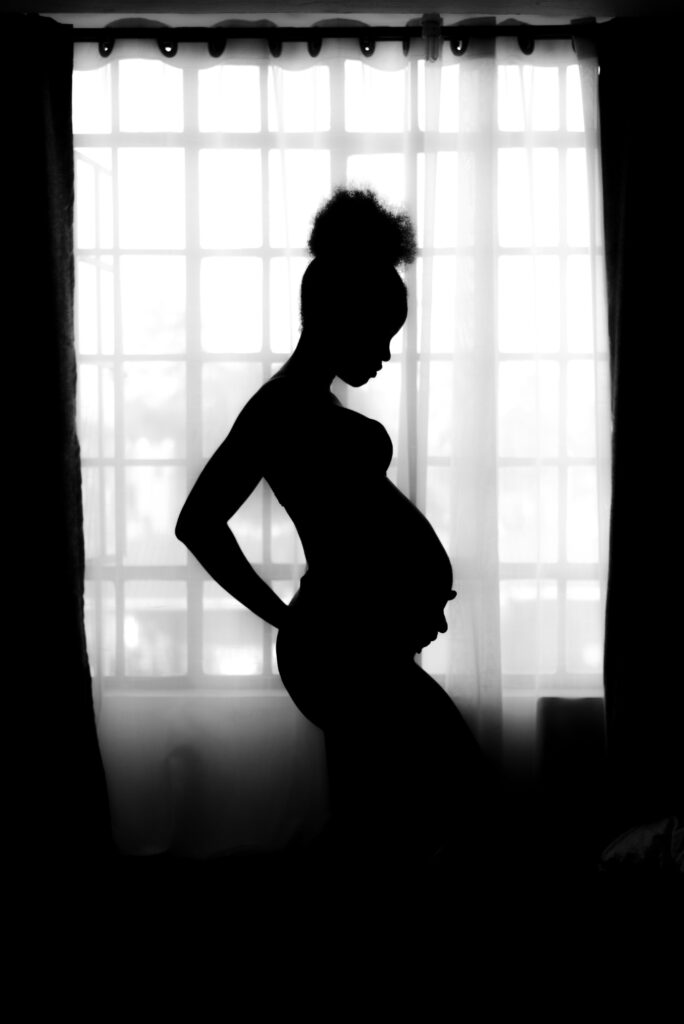 A black and white photo of an African-American pregnant woman holding her stomach in front of a window.