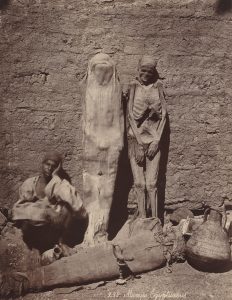 Image of a man selling mummies on the street. He is seated and two mummies are leant up against the wall.