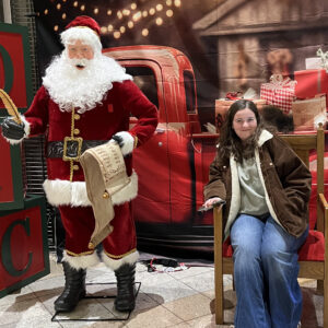 Mary sits in a chair next to a statue of Santa, smiling at the camera.