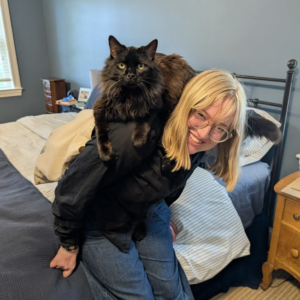 Eliza sits on a bed, smiling at the camera, with a large black cat sitting on her shoulders.