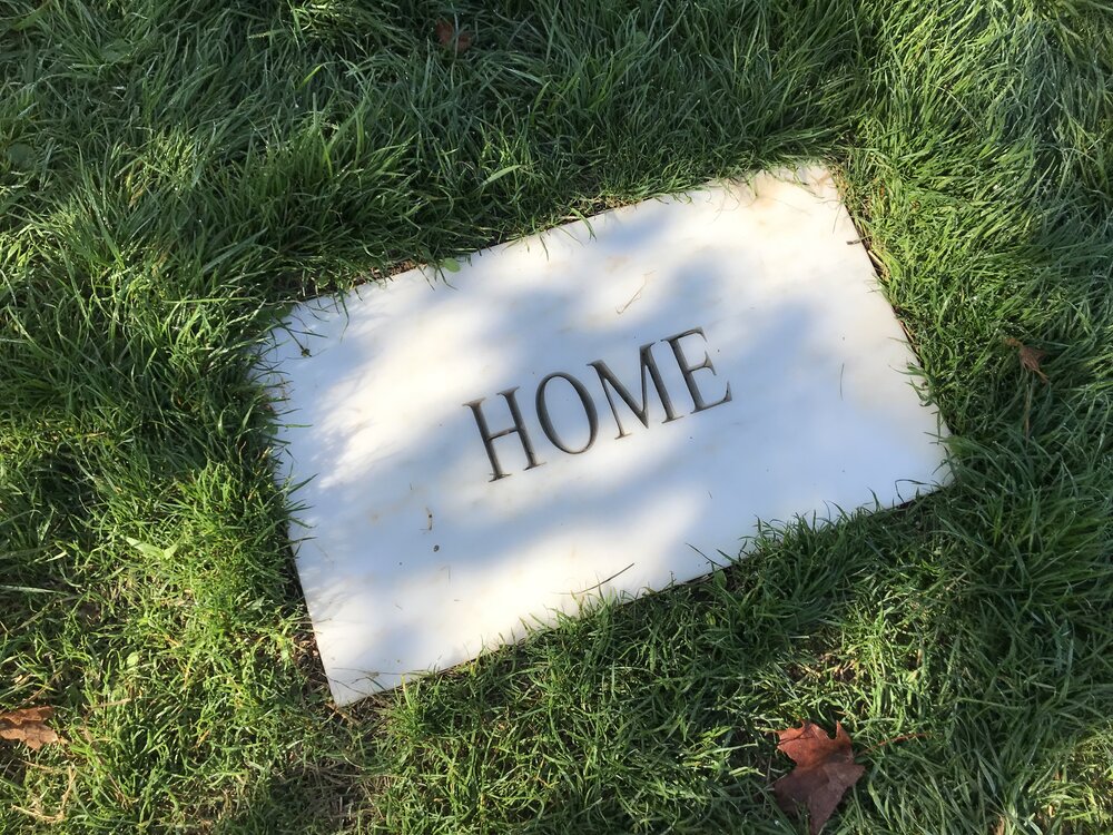 Close up of white marble stone flat against the ground with the word "HOME," in a grassy field
