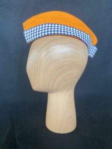 An orange hat with a blue gingham ribbon on a wooden mannequin head.