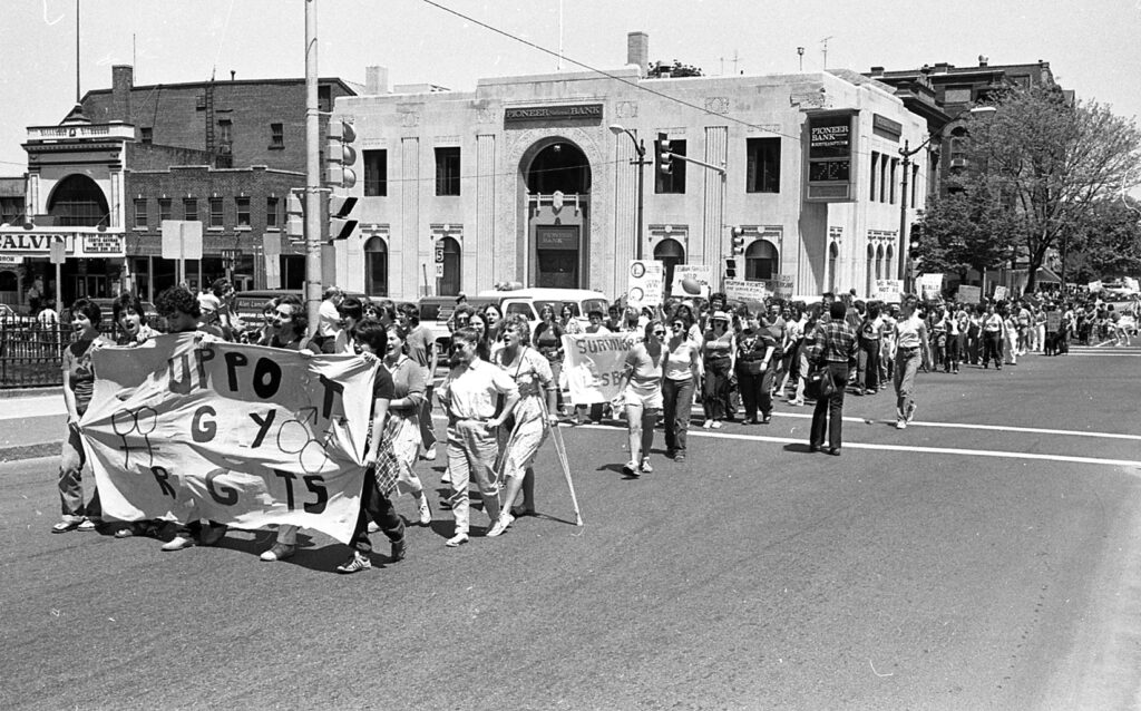 The Pride March travels down Main Street, crossing the intersection of Pleasant and Main. Participants are holding up signs, specifically a larger banner reading “Support Gay Rights.”