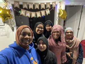 Six hijabi women joyously gathered with big smiles under a banner that reads "Ramadan Kareem" (generous Ramadan in English)