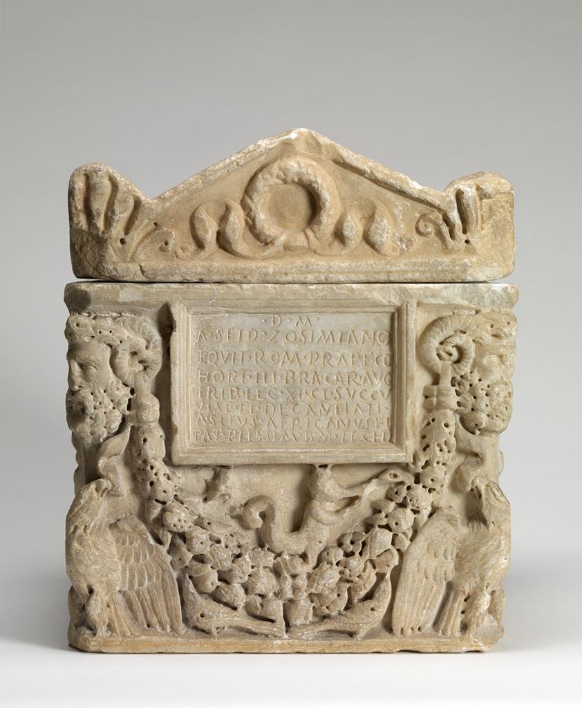 A carved stone box with images of faces on the corner and a plaque with words in the center.