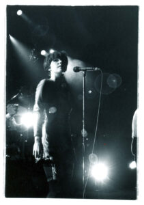 Black and white photo of Bjork performing on stage, standing at a microphone. Dramatic lighting creates lens flares, evoking a moody and atmospheric tone.