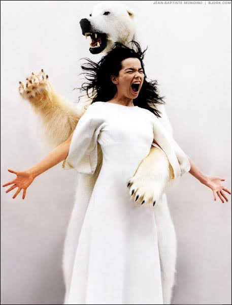 Bjork in a white dress screams with arms outstretched, as a polar bear stands behind her, paws wrapped around her shoulders. The scene is dramatic and surreal.