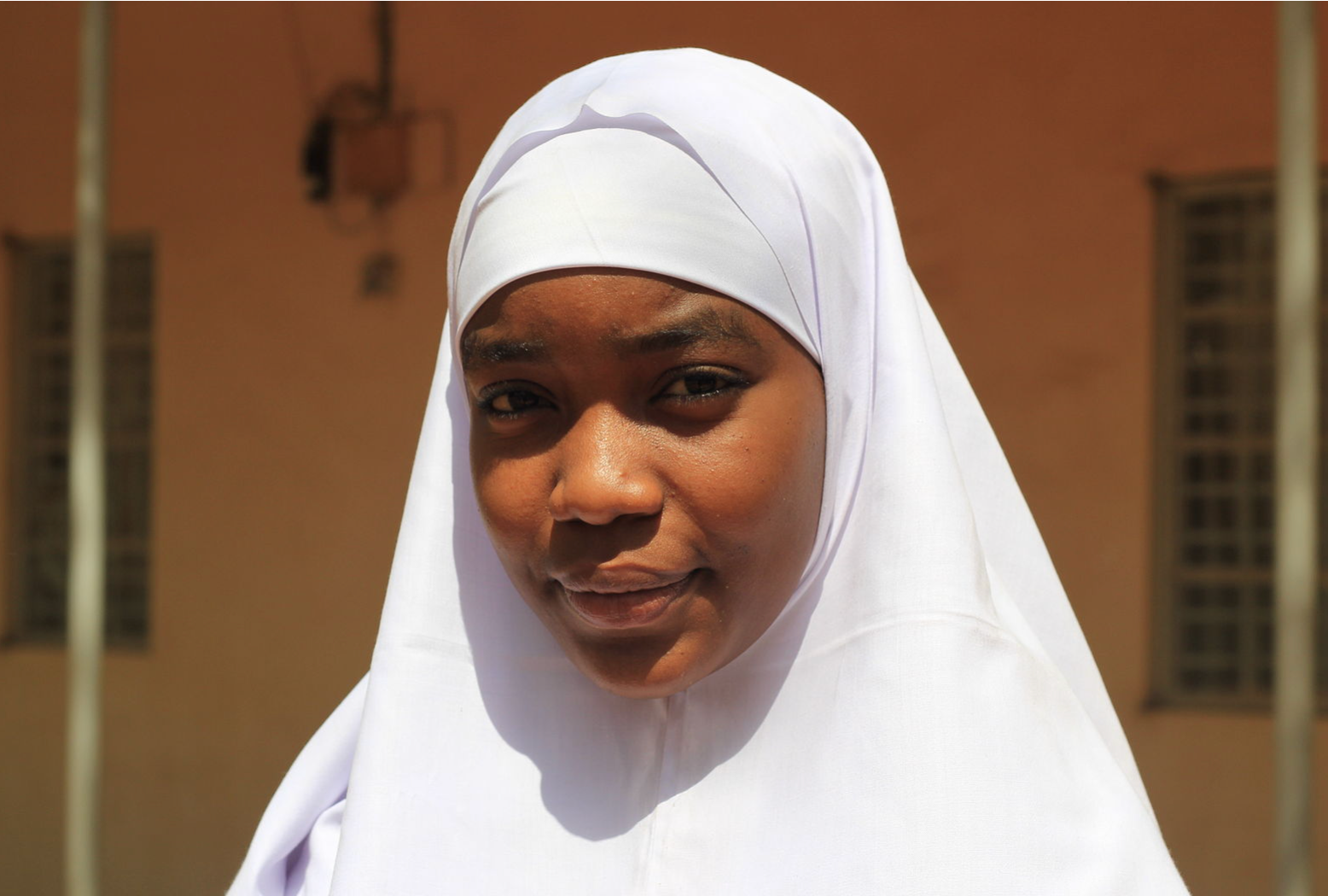 A young woman with a white hijabstands against a neutral background.