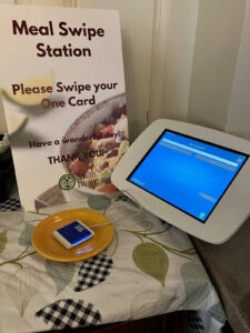 A meal swipe station with a sign reading "Please Swipe your One Card" and "Smith Dining." Nearby is a touchscreen and card reader on a table.