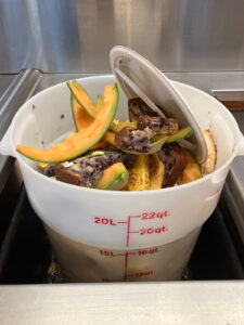 A large white container filled with food waste for composting, including melon rinds, banana peels, and cake pieces.