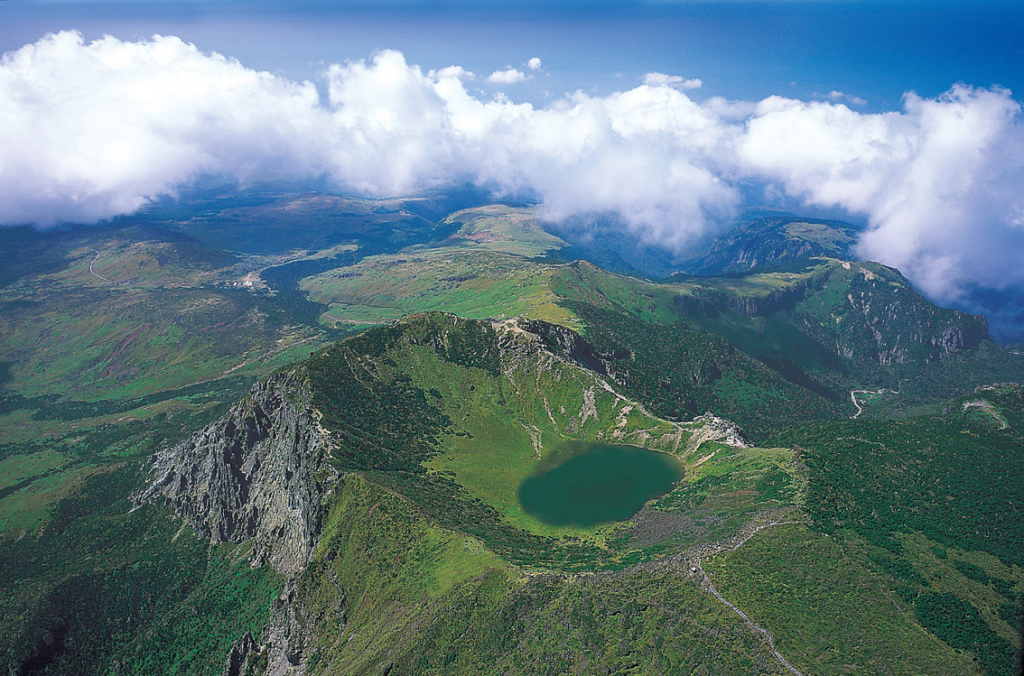 Aerial view of a lush green mountain landscape with a circular crater lake, surrounded by rocky peaks and soft clouds. The scene conveys serenity and majesty.