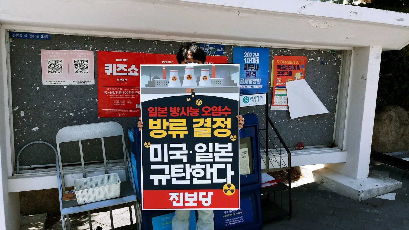 A person holds a protest sign in front of a bulletin board covered with colorful posters. The sign criticizesthe dumping of nuclear waste in South Korea. 