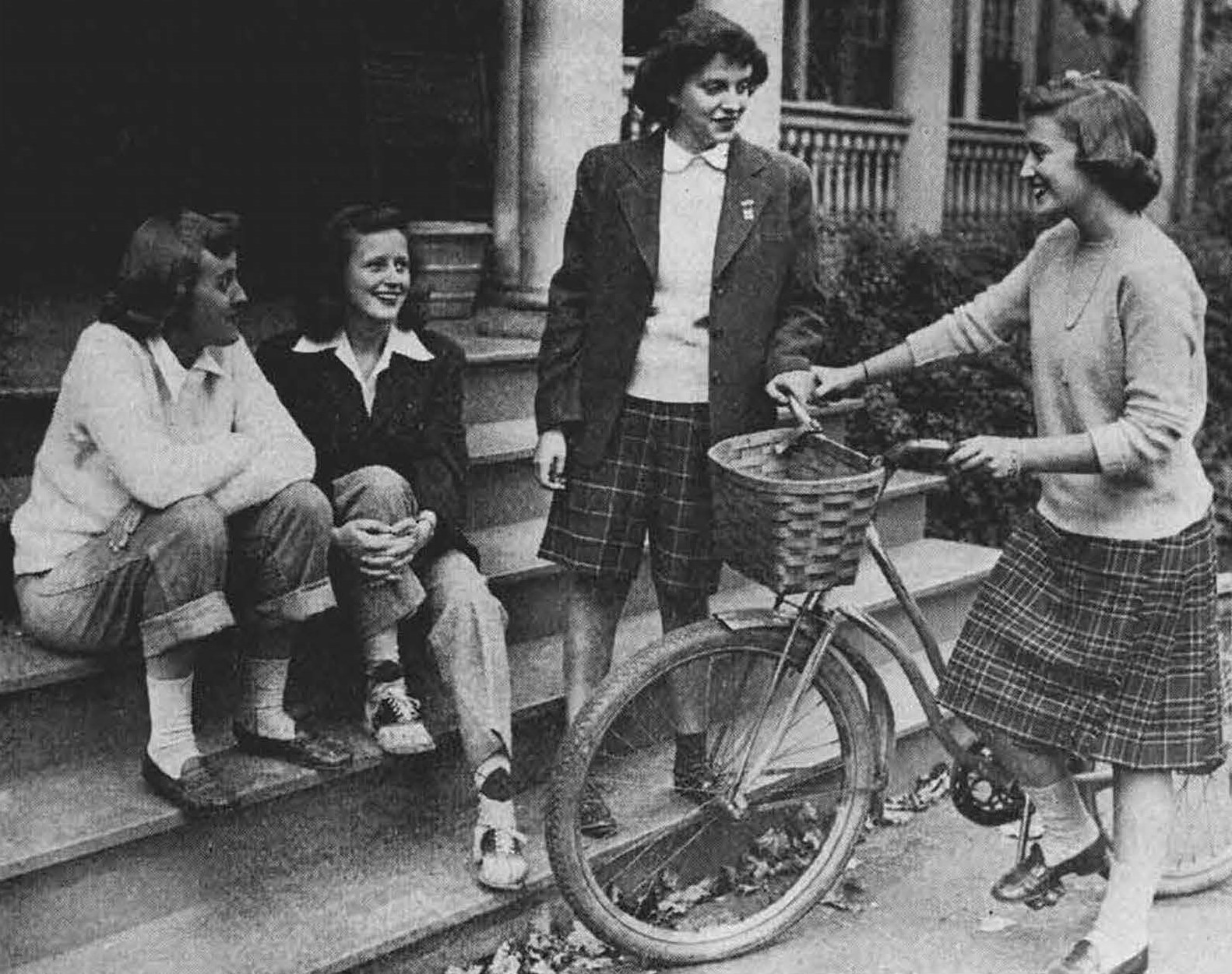 Black and white photo of four students, one in capris, one in slacks, one in shorts, and one in a skirt with a bicycle. They are smiling.