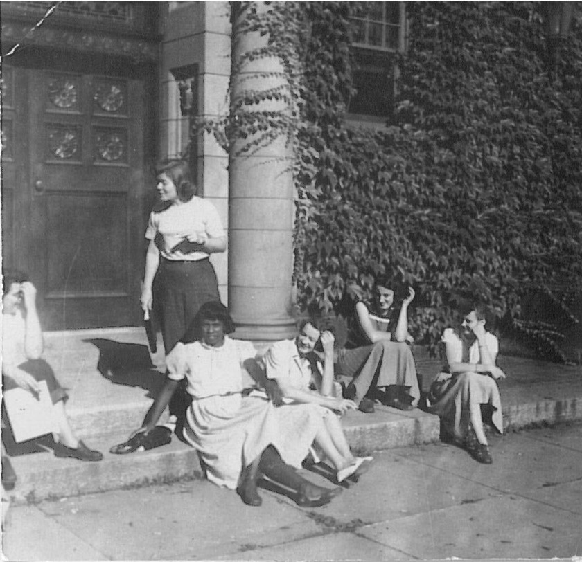 black and white photo of four people sitting in skirts and one person in pants standing.