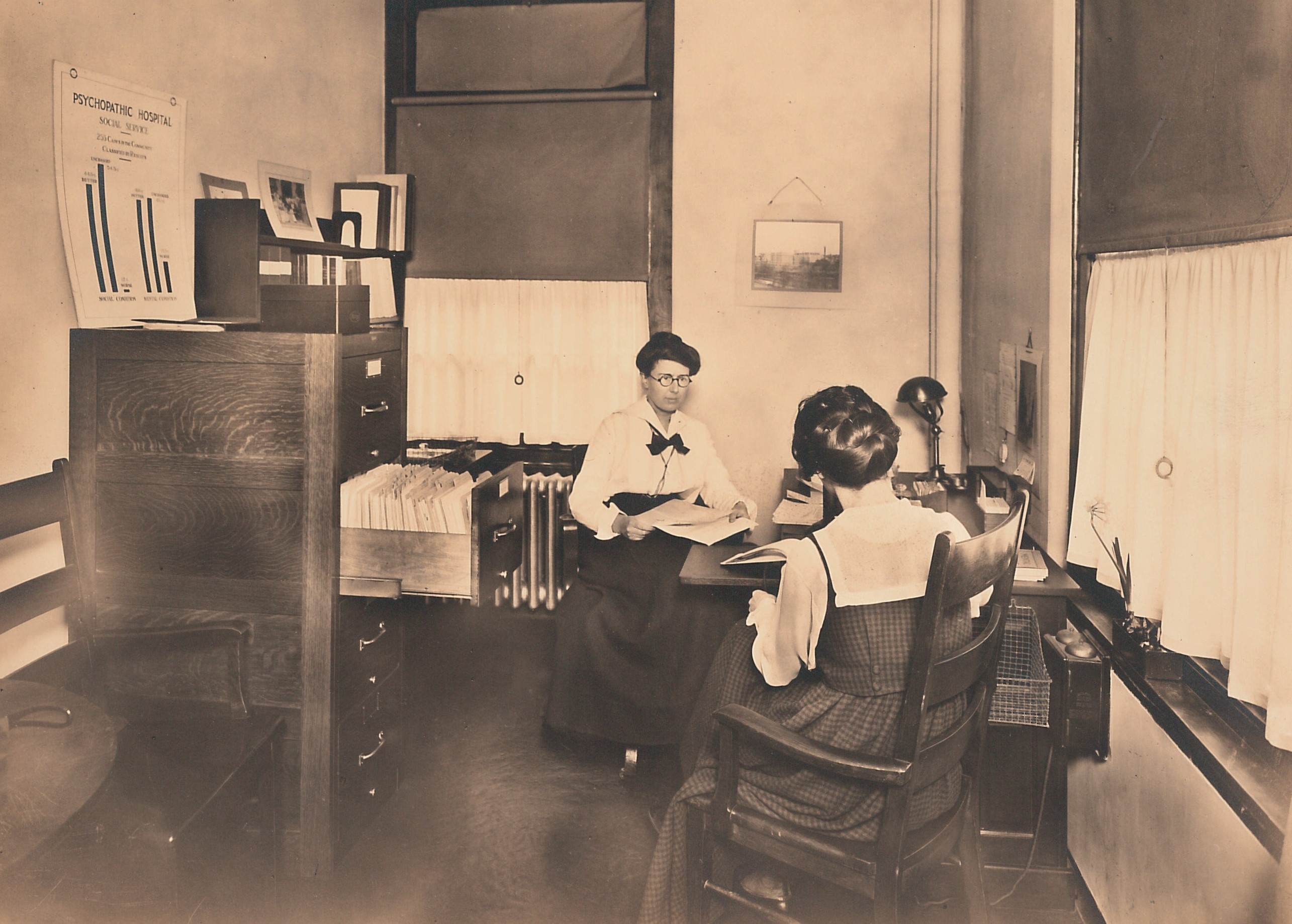 Black and white photo of Mary Jarrett in white blouse and black skirt at a desk with papers, sitting across from someone in a black dress with white blouse.