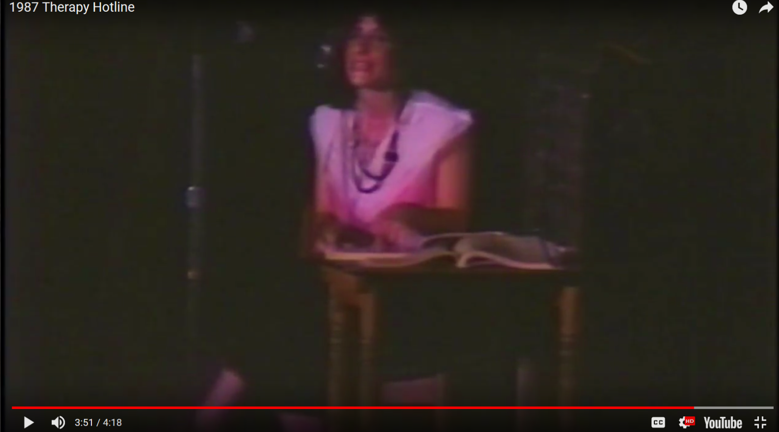 Screenshot of a youtube video. Dark stage with seated figure in pink light.