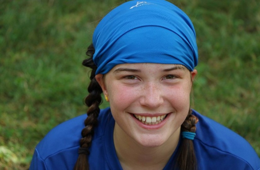 Image ID: a photo of Reed from the shoulders up wearing a blue atheletic headband that covers most of their head. They are sitting on the ground and smiling up at the camera.