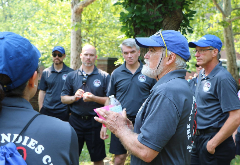 Image ID: several adults standing in a semi circle on a road lined by trees. They are all wearing black shorts, grey shirts, and many are wearing blue baseball caps. The focus of the photo is David Sloatman. He is holding a water bottle and speaking to the rest of the group.
