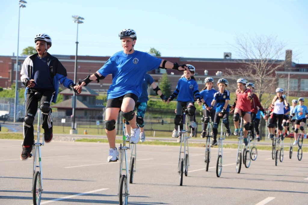 Image ID: the giraffe unicyclists of the Gym Dandies practicing their formations on a bright summer morning in a big parking lot. They are all in athletic gear and helmets riding in a big line while holding hands with their riding partners.