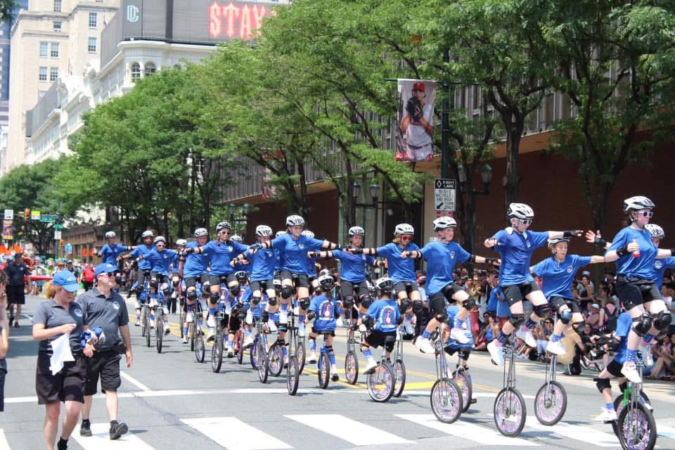 Image ID: a photo of the Gym Dandies performing a shoot the tunnel maneuver. Each Gym Dandie is wearing a blue shirt and black shorts. About 18 giraffe unicyclists are in a long line of groups of two and holding hands while ten 20 inch unicyclists ride in a straight line through the "tunnel" created by the giraffes. Parade viewers watch on both sides of the road.