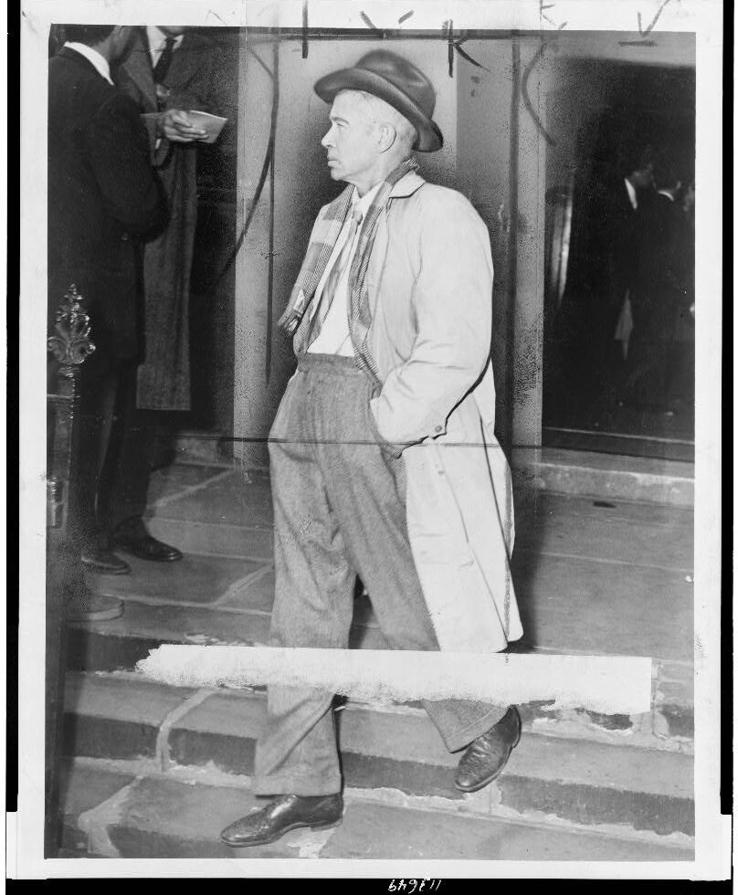 Black and white photograph of E.E. Cummings leaving a building---he is dressed smartly in some woven fabric trousers, a tartan scarf, and a hat.